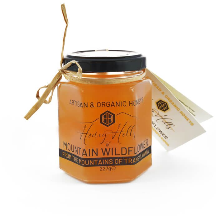 WildflowerTrans - Honey Hills - Online artisan shop with delicious, rare raw mountain honey varieties A hexagonal glass jar of Honey Hills Mountain Wildflower honey with a black lid, a raffia tie, and a tag. The label notes it is artisan and organic, from Transylvania.