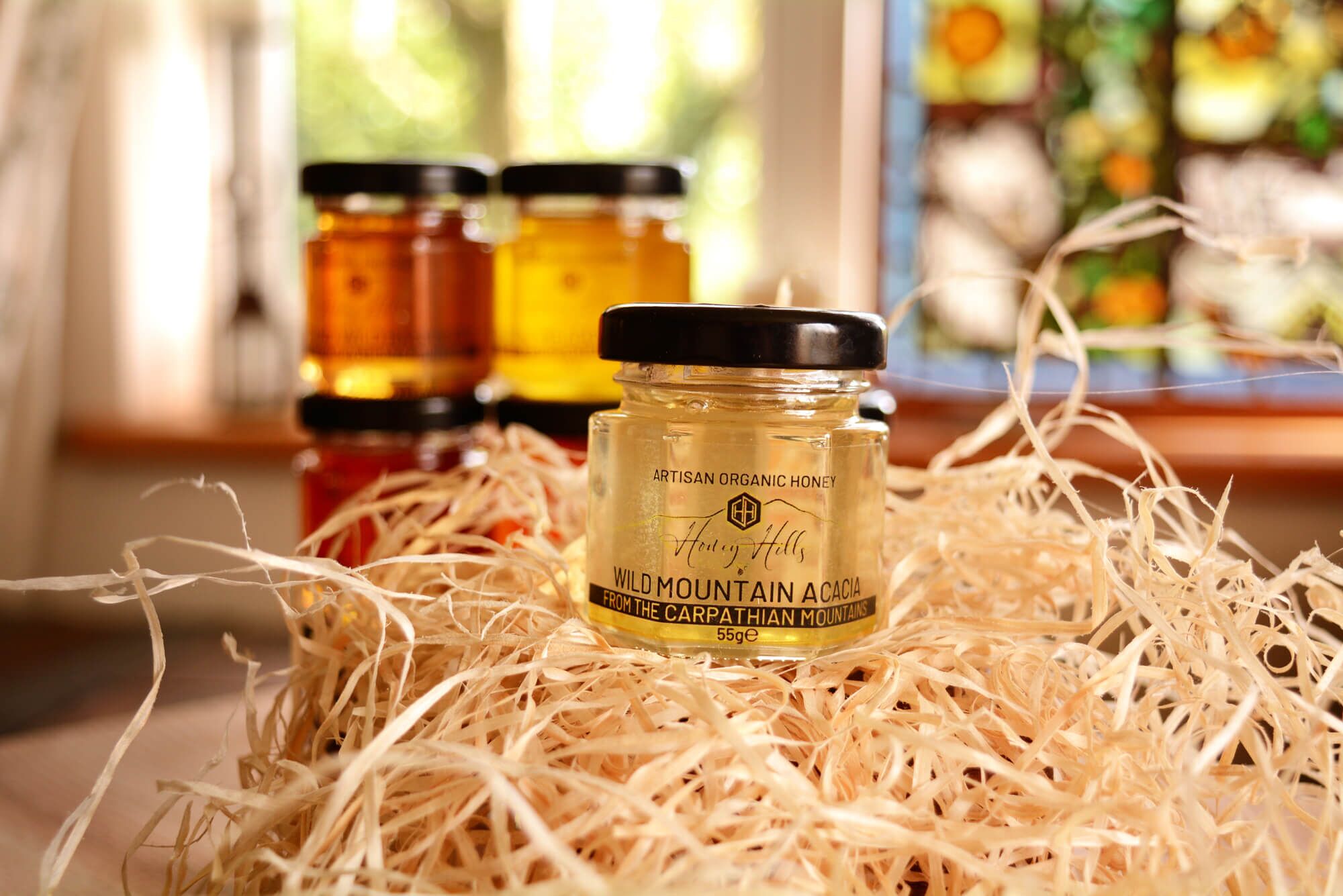 hamper-box-taster-set-acacia-honey - Honey Hills - Online artisan shop with delicious, rare raw mountain honey varieties Artisan organic honey jar labeled 'Wild Mountain Acacia' with colourful honey jars in the background on a rustic tabletop.